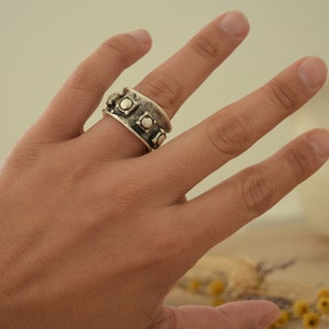 May include: A silver ring with a wide band and a row of raised squares with small circles in the center.