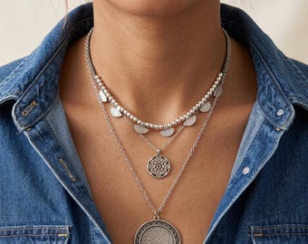 May include: A layered silver-tone necklace set featuring three distinct chains. The top layer has small metallic beads and semi-circular charms. The middle chain holds a circular pendant with a cut-out geometric pattern. The longest chain features a large, textured circular medallion with a decorative border.