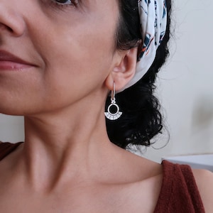 May include: Silver dangle earrings with a circle design and a small fan-shaped detail. The earrings are worn by a person with dark hair.