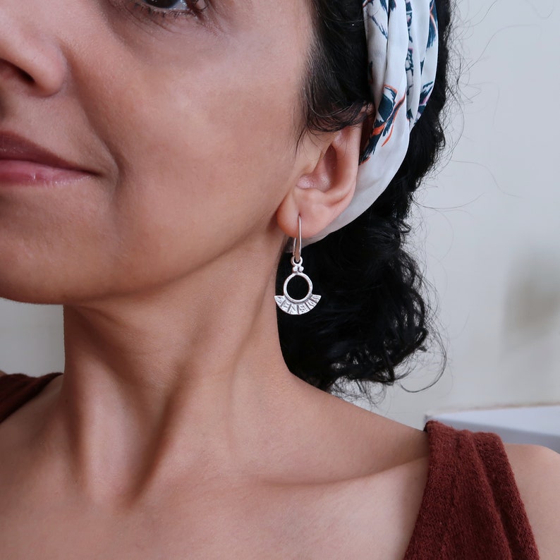 May include: Silver dangle earrings with a circle design and a small fan-shaped detail. The earrings are worn by a person with dark hair.