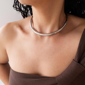 Könnte beinhalten: Eine silberfarbene Choker-Halskette. Die Halskette ist ein glattes, gebogenes Rohr, das eng am Hals anliegt. Das Model trägt ein braunes One-Shoulder-Top.