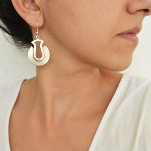May include: Silver hoop earrings with a unique cutout design. The earrings are worn by a person with dark hair.