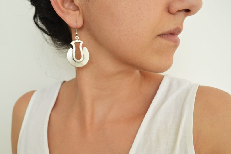 May include: Silver hoop earrings with a unique cutout design. The earrings are worn by a person with dark hair.