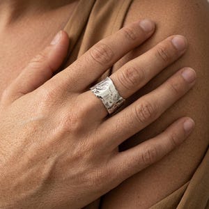 May include: A wide silver ring with a textured, uneven surface and a metallic finish. The band features an irregular, organic shape with a hammered appearance, wrapping around the finger with a distinct, rugged edge and a reflective, polished sheen.