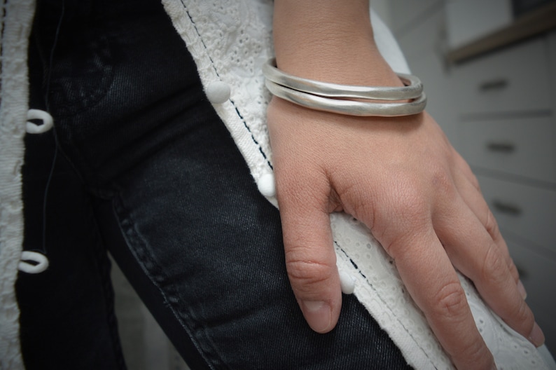 Op de afbeelding: Twee zilveren armbanden die om een pols worden gedragen. De armbanden zijn eenvoudig en glad, met een licht gehammerde textuur.