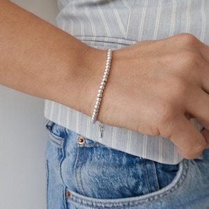 Puede incluir: Una pulsera de cuentas plateadas con un pequeño dije se usa en la muñeca. La pulsera está hecha de pequeñas cuentas redondas. La persona lleva un chaleco a rayas azul claro y vaqueros azules.