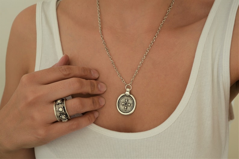 May include: A silver necklace with a round pendant featuring a cross design. The pendant has a textured surface and is attached to a silver chain. The necklace is worn by a person wearing a white tank top.