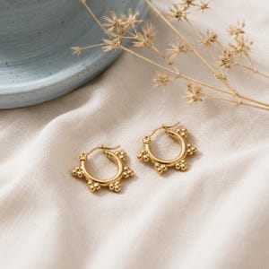 Könnte beinhalten: Ein Paar goldene Creolen, bestehend aus einem kreisförmigen Ring, der am äußeren Rand mit kleinen, gleichmäßig verteilten kugelförmigen Perlen verziert ist. Die Ohrringe haben eine polierte metallische Oberfläche und einen sichtbaren Klappverschluss.