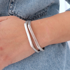 May include: Two silver bracelets stacked on a person's wrist. The bracelets are made of a smooth, flat metal and have a simple, modern design.