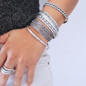 May include: A collection of silver bracelets and rings. The bracelets feature various designs, including braided, patterned, and beaded styles. The rings have a stacked design. The jewelry is worn on a person's wrist and hand.