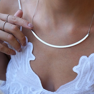 May include: A silver necklace with a thin, curved bar pendant. The necklace is worn by a person with a white top.