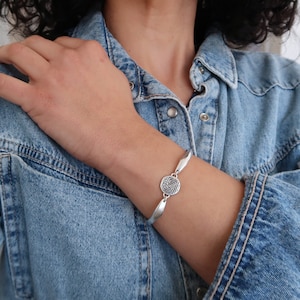 Könnte beinhalten: Ein silbernes Armband mit einem runden Anhänger, der ein geometrisches Labyrinth-Design zeigt.