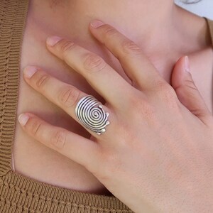 Op de afbeelding: Een zilveren ring met een spiraalvormig ontwerp. De ring wordt gedragen aan de wijsvinger van een hand.