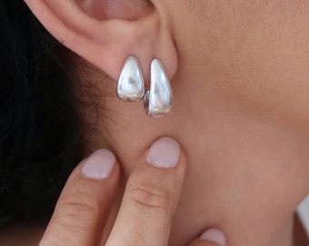 May include: A pair of small, polished silver-tone hoop earrings with a smooth, teardrop-shaped design. The earrings feature a reflective metallic finish and a secure hinged closure, resting against the earlobe in a simple, curved silhouette.