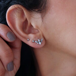 May include: A close-up of a person's ear with three silver earrings. The earrings are small and simple, with one featuring a bee design.