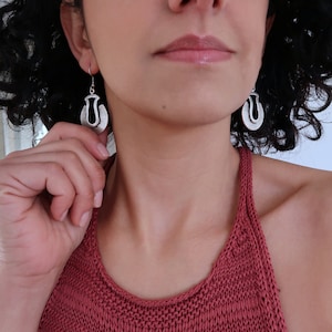 May include: A woman wearing a red knitted tank top with silver earrings shaped like a stylized letter U.