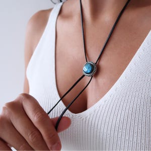 May include: A bolo tie with a round, iridescent blue stone pendant and a silver-coloured beaded border. The pendant hangs from a black cord, a stylish and unique accessory.