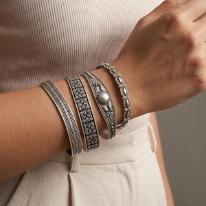 May include: A set of four silver-toned metal cuff bracelets. One features a twisted rope pattern, another has a repeating square floral motif, the third displays a central circular bead with leaf accents, and the fourth has a linked oval design.