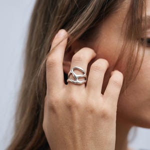 Könnte beinhalten: Ein silberfarbener Ring mit einem offenen, fließenden und organischen Ausschnitt-Design mit glatten, abgerundeten Kanten. Das Band hat eine durchgehende, unregelmäßige Form, die den Finger umschließt und eine Reihe verbundener Hohlräume bildet.