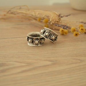 May include: Two silver rings with a decorative design of raised circles around the band. The rings are on a light brown wooden surface.