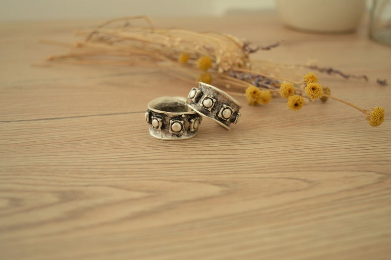 May include: Two silver rings with a decorative design of raised circles around the band. The rings are on a light brown wooden surface.