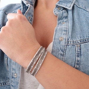 Puede incluir: Un conjunto de cinco pulseras de plata con un patrón geométrico. Las pulseras están apiladas en la muñeca de una persona.