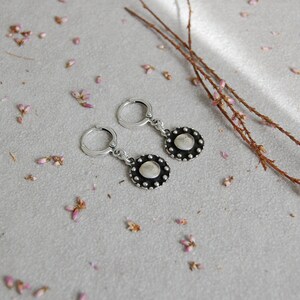 May include: A pair of silver hoop earrings with a round, silver charm that has a black center and a circle of small silver beads around the edge.
