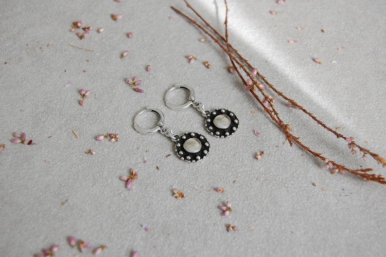 May include: A pair of silver hoop earrings with a round, silver charm that has a black center and a circle of small silver beads around the edge.
