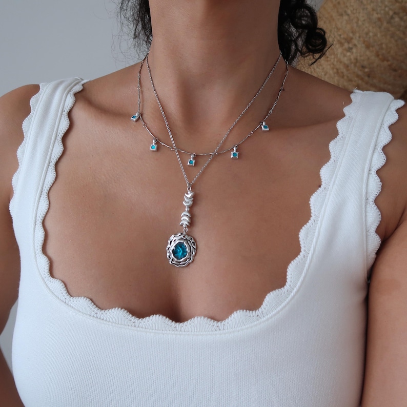 May include: A silver necklace with a turquoise pendant featuring a circular design. The pendant is surrounded by smaller turquoise stones. The necklace is layered with a delicate chain with small turquoise stones hanging from it.