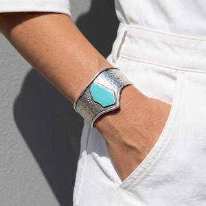 May include: A wide cuff bracelet featuring a textured, hammered silver-tone metal finish and a central, smooth, geometric turquoise-coloured inlay. The bracelet has a contoured shape that widens towards the centre, showcasing a polished metallic edge.