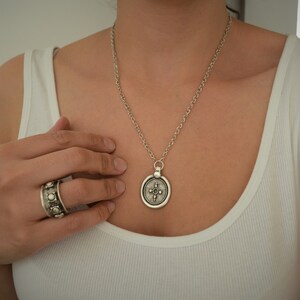 May include: A silver necklace with a round pendant featuring a detailed design. The pendant has a cross-like shape with a circle in the center. The necklace is worn by a person with a white tank top.