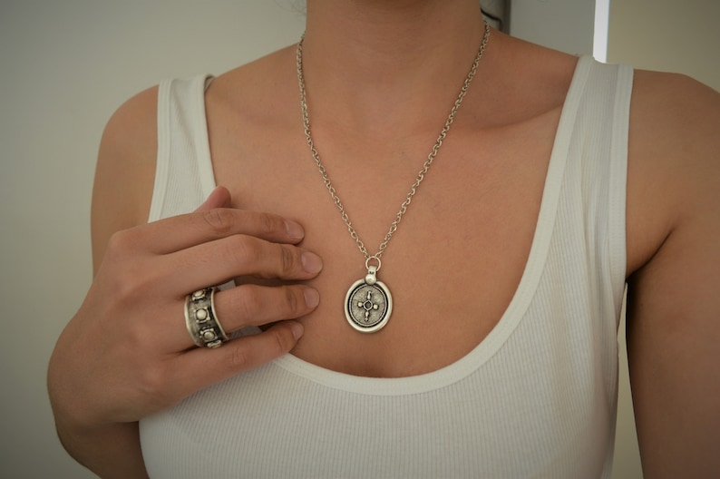 May include: A silver necklace with a round pendant featuring a detailed design. The pendant has a cross-like shape with a circle in the center. The necklace is worn by a person with a white tank top.