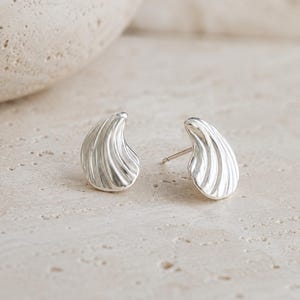 May include: A pair of silver stud earrings featuring a curved, teardrop-like shape with vertical ribbed detailing. The earrings have a polished, reflective metallic finish and include a standard post backing for secure attachment to the earlobe.