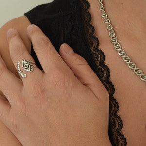 May include: A silver eye ring with a detailed design. The ring is worn on the middle finger of a person's hand. The person is wearing a black lace top and a silver chain necklace with a large crystal pendant.