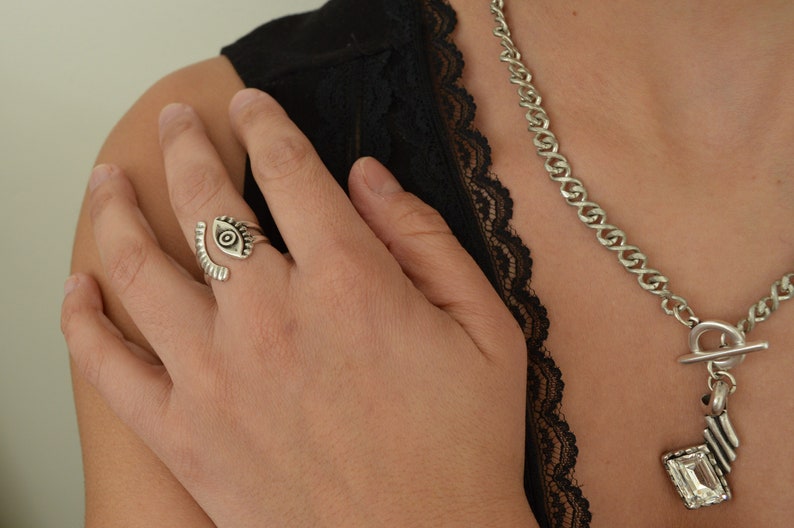 May include: A silver eye ring with a detailed design. The ring is worn on the middle finger of a person's hand. The person is wearing a black lace top and a silver chain necklace with a large crystal pendant.