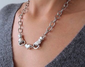 May include: A silver chain necklace with two hand-shaped charms and a heart charm in the center.