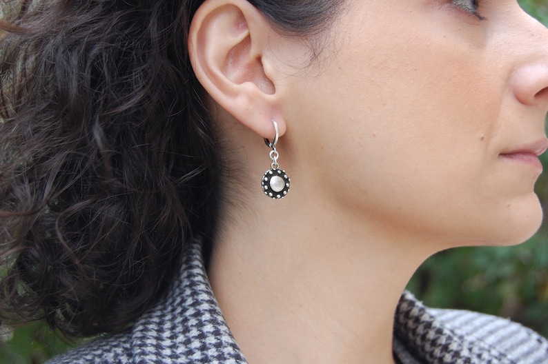 May include: Silver hoop earrings with a round, silver charm featuring a textured, circular design with a raised center.