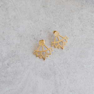 May include: A pair of gold-colored earrings with a geometric, fan-like design. Each earring features a small, round stud with linear detailing, connected to a larger, openwork shape. The earrings are displayed on a light gray surface.