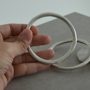 Op de afbeelding: Twee zilveren armbanden, een groter dan de andere, liggen op elkaar. De armbanden zijn glad en hebben een afgeronde vorm.