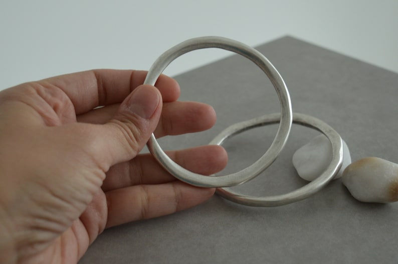 Op de afbeelding: Twee zilveren armbanden, een groter dan de andere, liggen op elkaar. De armbanden zijn glad en hebben een afgeronde vorm.