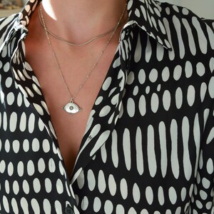 May include: A silver necklace with a small eye-shaped pendant hangs on a person wearing a black and white polka dot shirt.