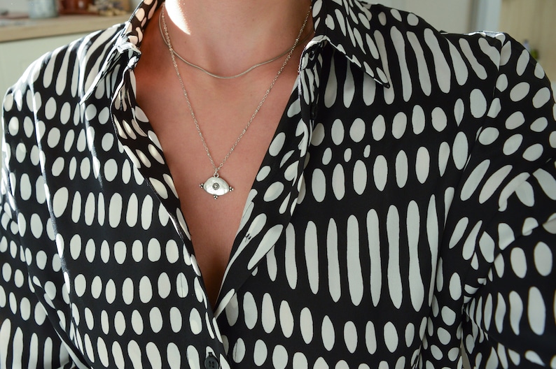 May include: A silver necklace with a small eye-shaped pendant hangs on a person wearing a black and white polka dot shirt.