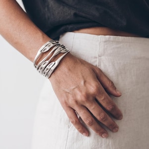 May include: A wide silver-toned cuff bracelet featuring multiple interconnected, curved metal bands with a textured, hammered finish. The bracelet wraps around the wrist, creating an open, layered structure with smooth, rounded edges and a polished metallic sheen.