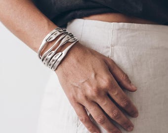 Puede incluir: Una pulsera brazalete ancha de tono plateado que presenta múltiples bandas metálicas curvas e interconectadas con un acabado texturizado y martillado. La pulsera envuelve la muñeca, creando una estructura abierta y superpuesta con bordes suaves.