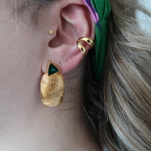 Könnte beinhalten: Goldene Ohrringe mit einem strukturierten, fingerabdruckartigen Design und einem dreieckigen grünen Stein. Ein goldener Ohrklemmen und ein kleiner sternförmiger Ohrstecker sind ebenfalls zu sehen. Ein auffälliges Schmuckstück.