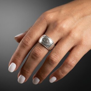 Peut inclure: Une large bague en argent avec une texture martel&eacute;e et un symbole d'&oelig;il protecteur grav&eacute; au centre. Le m&eacute;tal pr&eacute;sente une finition polie avec des d&eacute;tails assombris &agrave; l'int&eacute;rieur du motif de l'&oelig;il, cr&eacute;ant un contraste net sur la surface.