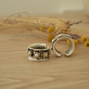 May include: Two silver rings with decorative details. The rings are on a wooden surface with dried flowers in the background.