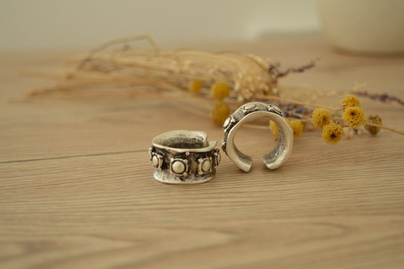 May include: Two silver rings with decorative details. The rings are on a wooden surface with dried flowers in the background.