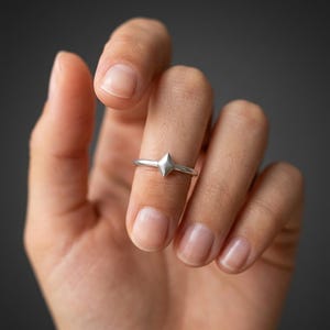 May include: A silver ring featuring a small, faceted diamond-shaped centerpiece. The band is thin and smooth, with a polished metallic finish. The geometric focal point sits centered on the band, creating a simple and clean aesthetic for the finger.