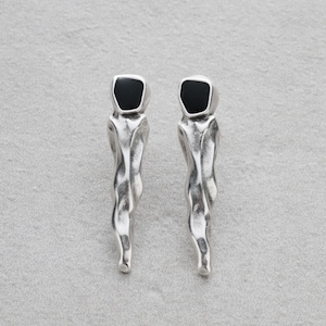 Puede incluir: Un par de pendientes plateados con un diseño único. Cada pendiente presenta una parte superior negra de forma cuadrada y un cuerpo alargado texturizado. Los pendientes tienen una estética moderna y abstracta.